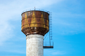White with yellow brown color rusted old water tower on the background of a bright blue spring sky. With cornice and stairs.