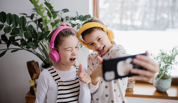 Two small girls sisters with headphones indoors at home, grimacing when taking selfie.