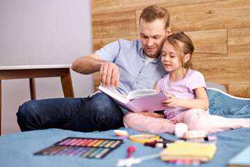 Obraz na płótnie Canvas young father explaining paragraph presented in textbook to his little daughter, she listening to him with interest, sitting on bed at home. family concept