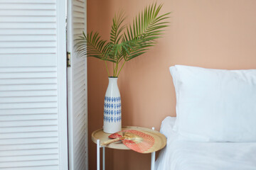 Cozy interior of the white retro wardrobe, white bed and wooden table and white vase on it with green plants on the background of the peach wall