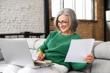Happy mature businesswoman accountant in glasses and green jumper sitting on the couch with laptop on her lap, taking notes, holding papers, contract studying or working remotely online from home