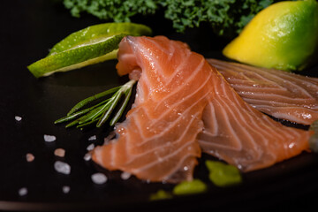 Three Salmon Slices Served On A Black Ceramic Plate With Kale Leaf, Avocado, Lime Wedges, Rosemary Sprig And Coarse Tibetan Salt. Organic Food. Side View. Serving Example. Selective Focus.