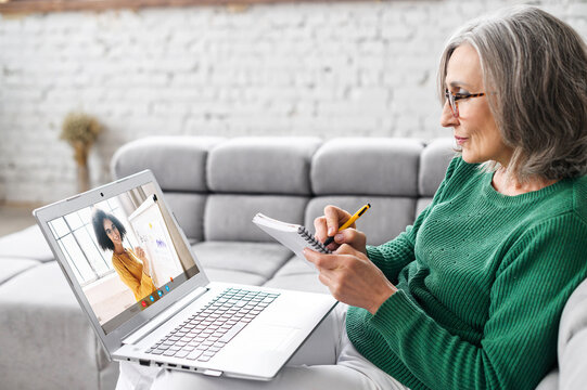 Attentive Mature Gray-haired Businesswoman In Glasses Sitting On The Couch At Home, Studying, Taking Virtual Class, Having Online Meeting On A Laptop, Taking Notes And Listening To Female Teacher