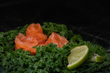Three Salmon Slices, Each Rolled Into A Rose, Served On A Kale Leaf, Garnished With Lime Wedges. Side View. Example Of Serving On A Black Ceramic Plate. Selective Focus. Total Black Back.