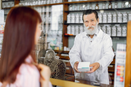 Smiling Friendly Senior Bearded Man Pharmacist, Standing At The Counter In Old Pharmacy, Offering Medicine To Young Female Patient. View Over The Clients Shoulder Of The Pharmacist