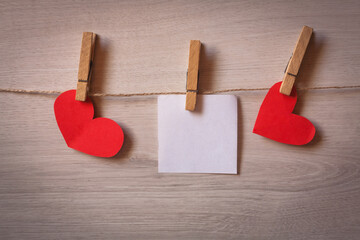 Clothespins with blank sheets of paper hanging on wooden background with hearts.