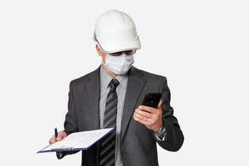 Man in suit, protective mask and white hardhat on his head holds clipboard and looks at smartphone screen. Isolated on light gray background