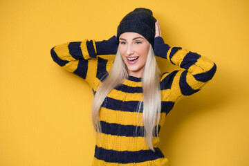 Shocked amazed woman, model wearing woolen cap and sweater, isolated on yellow background. Surprised girl with hands on head. Amazement concept