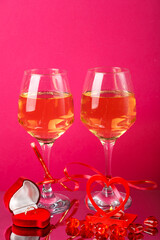 Two glasses of champagne with red ribbons on a pink background next to a heart candlestick with a burning candle and a box ring.