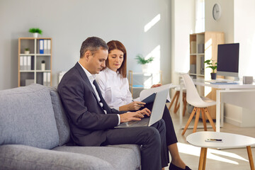 Obraz premium Two serious focused business people sitting on couch while meeting in the office. Man and woman working with data together, using laptop computer, doing paperwork