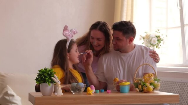 Happy easter. Family with faces stained with paint painting eggs at home, having fun. Father, mother and daughter wearing bunny ears