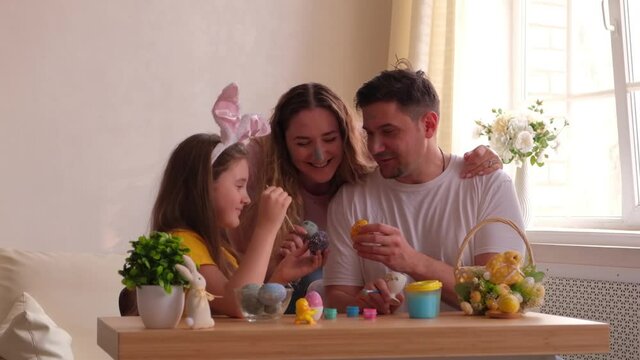 Happy easter. Family with faces stained with paint painting eggs at home, having fun. Father, mother and daughter wearing bunny ears