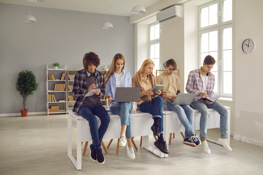 Group Of Classmates Getting Ready For Examination. Busy College Students Sitting On Classroom Desk With Modern Laptops And Tablets, Studying Course Books, Looking Through Lecture Notes, Doing Homework