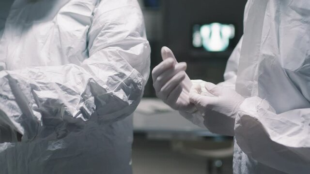 Midsection close up with slow motion of two unrecognizable doctors in dark hospital room wearing protective white costumes and gloves