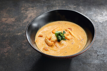An oriental soup with tomatoes, coconut milk, shrimps, bamboo shoots, on a black stone background.
