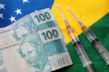 Brazilian money and syringes on top of Brazilian flag. Vaccination against COVID-19 in Brazil. Vaccines prices concept.
