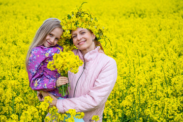 Fototapeta premium Cute girl with long light hair hug her mother in the field of yellow flowers. Rural landscape of blooming rapeseed. Mother's day concept.
