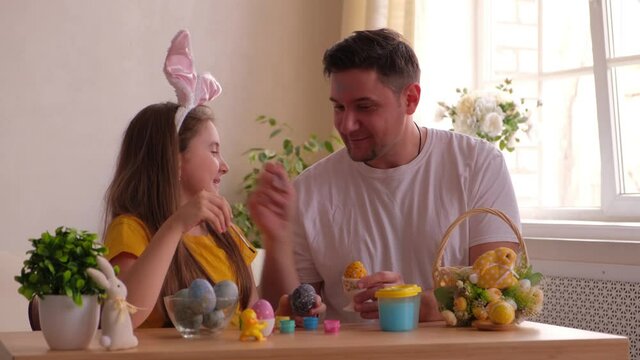 dad and daughter stain each other's faces with blue paint for painting eggs. on the table is a basket with Easter eggs and paints.