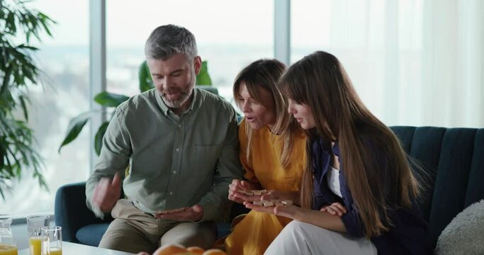 Excited caucasian family trio of parents couple with daughter playing domino table game inside apartment. Home leisure. Lockdown. Holidays.