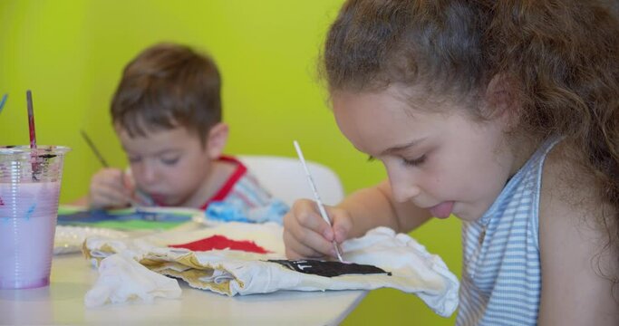 Happy cute children, a girl of 7-8 years old and preschool boy, paint at home with watercolors sitting at a white table, little boy draws on paper with colored paints and baby girl on her white shorts