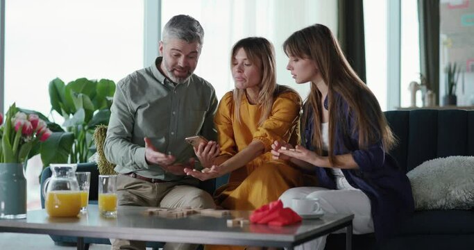 Cheerful parents couple with adult daughter playing table game with domino wooden sticks having fun watching tutorials on mobile phone internet.