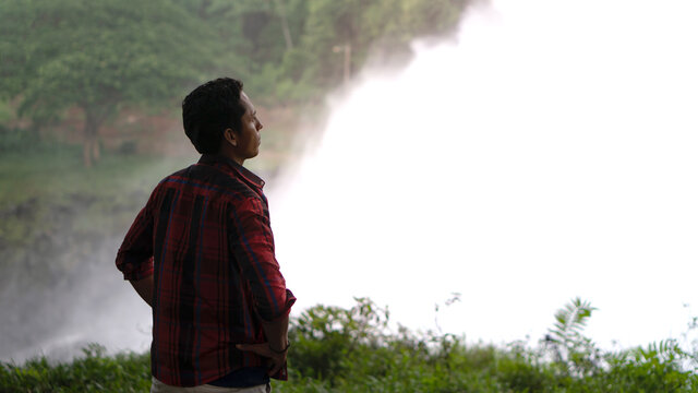 Backlit latin man on the bank of a river with waterfall, man wearing red t-shirt, horizontal and panoramic image - Powered by Adobe