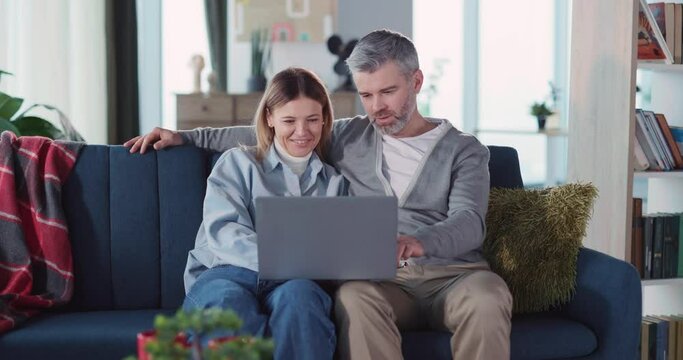 Caucasian casual adult couple of married man with woman using portable laptop pc computer watching movie film together in living room.