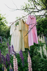 Natural colored dresses hanging on on a tree in the garden with lupine flowers. Concept organic clothes, eco-friendly, ecological fashion