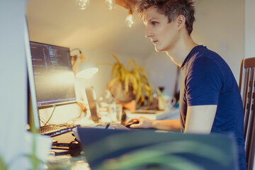 A man sitting at a desk working from home on his computer. IT specialist programmer writing code on his pc and laptop. Developer remote home office. Freelance workspace. Soft selective focus.