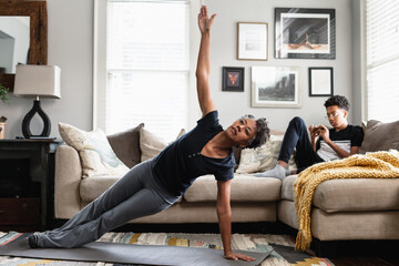 Black mom working out in family room, teenage son on phone in background, side plank exercise