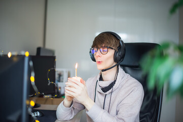 Young man blowing candle on the festive cupcake on virtual birthday call party during lockdown. Generation Z man celebrating his birthday alone on video call in his room. Social distance.
