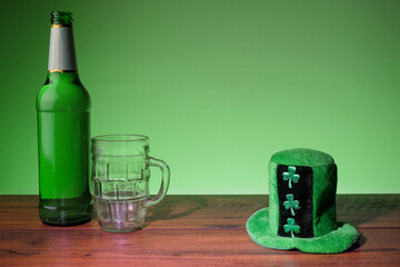 Green bottle of lager and glass and green hat with shamrock on a wooden bar surface. Green background. Saint Patrick theme background