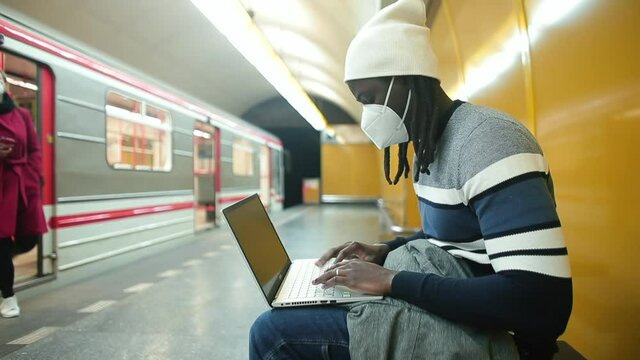 A black male freelancer wearing a mask uses a laptop at a train station. Route planning. Control of digital data in quarantine. Train in bokeh.