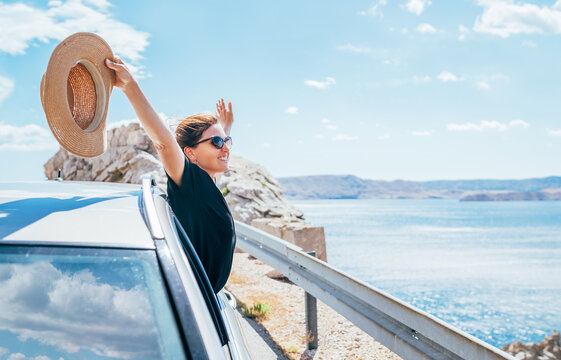 Cheerful Woman portrait enjoying the seaside road trip. Dressed a black dress with straw hat and sunglasses she wide opened arms and shining with happiness. Summer vacation traveling by auto concept.