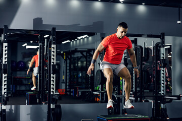 Fototapeta na wymiar A strong young man in sportswear and armband jumps on a stepper in an indoor modern gym with a mirror. Healthy lifestyle, fitness training