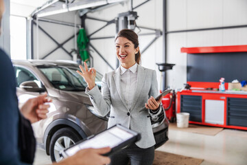 Smiling friendly car seller talking to a mechanic and showing okay hand gesture while standing in car salon.