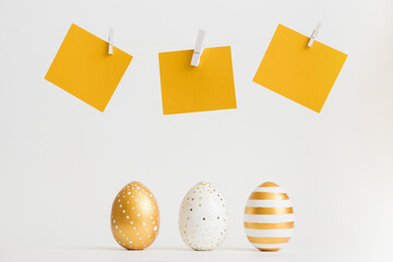 Three Easter golden decorated eggs stand in a row with yellow text stickers above them on white background. Minimal easter concept. Happy Easter card with copy space for text. Top view, flatlay