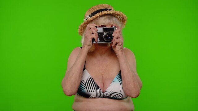 Travel, long awaited summer holiday vacation, beach party. Senior woman tourist photographer in swimsuit taking photos on old retro camera, smiling on chroma key. Grandmother relaxing on sea resort