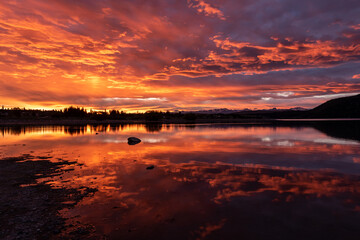 The shore and trees silhouetted against an intense, orange sunset and reflected in Lake Tekapo, Mackenzie District, Canterbury, New Zealand.