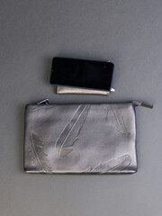 Silver leather handbag with embossed feathers. Women's cross body lies on a gray background with a wallet and phone.