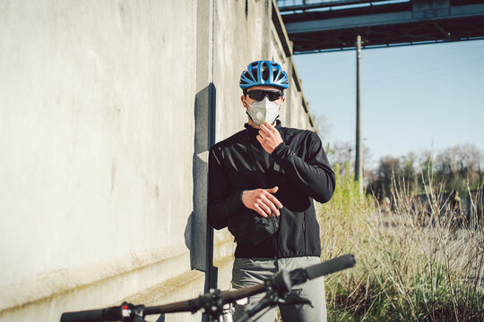 A man cyclist in a protective face mask stands next to a bicycle on a background of gray concrete wall. Unrecognizable Courier in respirator. Delivery service during a pandemic