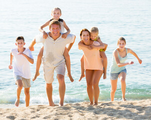 Smiling parents with four children in different ages sitting on parents back taking walk on beach
