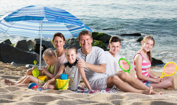 Carefree Family With Children Together On Beach Sitting Under Umbrella And Relaxing Weekend