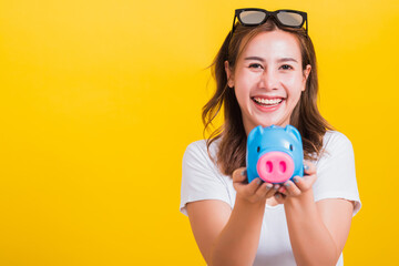 Portrait Asian Thai beautiful happy young woman smiling hold piggy bank with a lots money and looking to camera, studio shot isolated on yellow background, with copy space