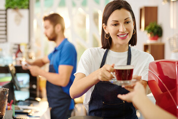 Pretty smiling female barista giving cup of fresh coffee to customer