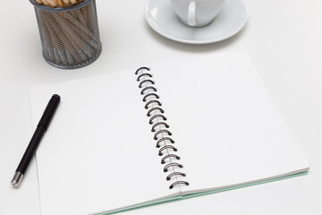 A blank notebook on a white table with pens, coffee mugs and pencils