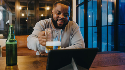 Middle-aged African American male drinking beer having fun happy night party event online celebration via video call in living room at home. Social distancing, quarantine for coronavirus prevention.