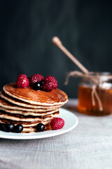 Juicy pancakes with berries and honey on a white plate, spoon, jar, wooden table