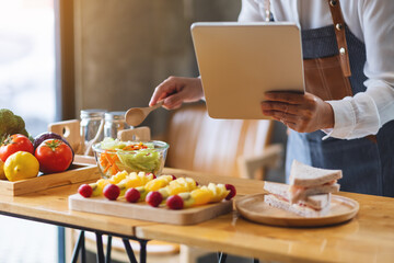 A woman following recipe on digital tablet while cooking salad and sandwich in the kitchen, online learning cooking class concept
