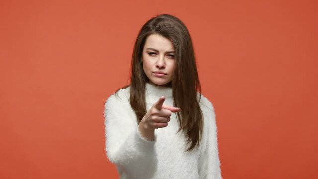 Are you ready? Serious bossy brunette woman in fluffy white sweater pointing finger at you looking at camera with confidence, making choice. Indoor studio shot isolated on orange background